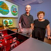 Martin Gruebele and Zaida Luthey-Schulten stand side-by-side next to lab equipment with illustrations of images on a screen on the wall behind them.