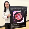 Xiaolin Liu stands holding a large poster of the winning Rose in a Flask image. 